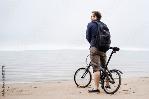 young caucasian man with a small backpack standing with the bike on the beach looking at the sea and preoccupied at the sight of worsening weather