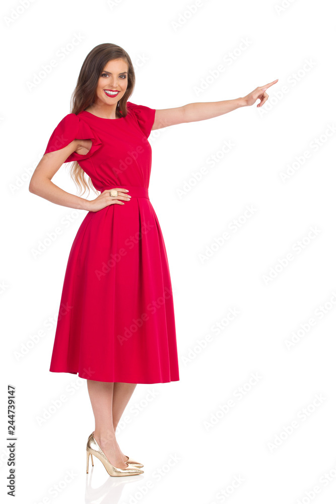 Fototapeta premium Uśmiechnięta młoda kobieta w czerwonej sukience i złotych szpilkach patrzy na aparat i wskazuje