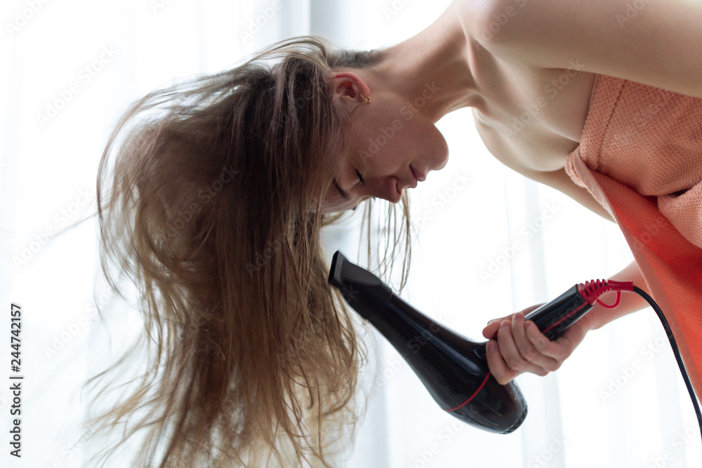 Fototapeta premium Beautiful young brown haired woman in bath towel is using a hair dryer for drying her long hair after shower in room. Hair Care.