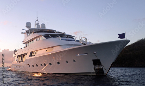 Slika na platnu A large, white luxury power yacht at anchor with its shiny hull reflecting the pink sky at sunset, floating on calm dark water with a baby blue to purple sky in the background