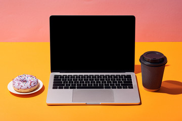 laptop with blank screen near donut and paper coffee cup on yellow surface and pink background