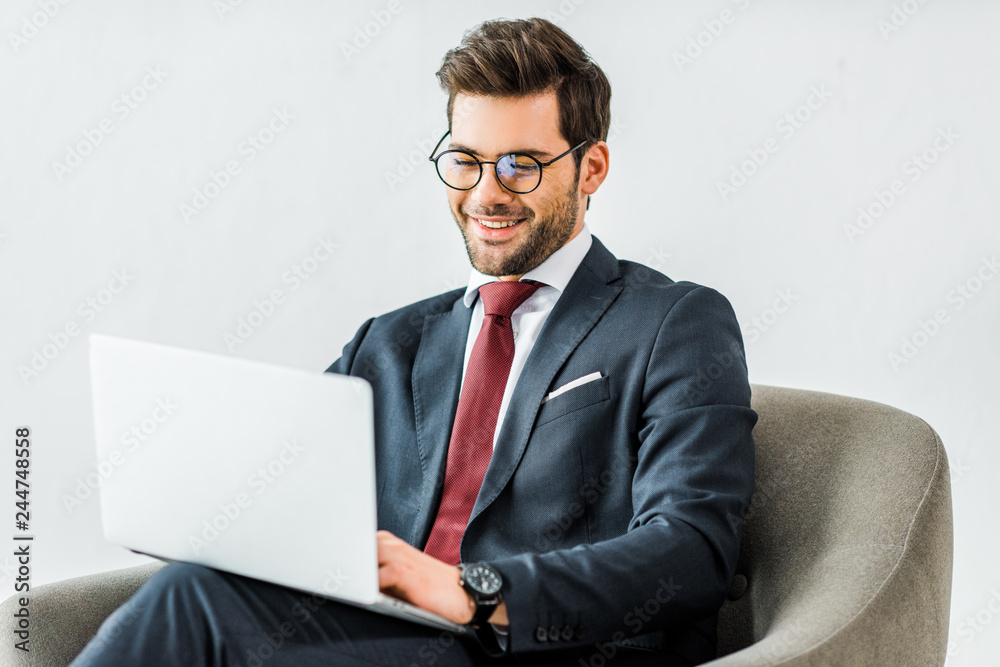 smiling businessman in formal wear sitting on armchair and using laptop in office smiling businessman in formal wear sitting on armchair and using laptop in office