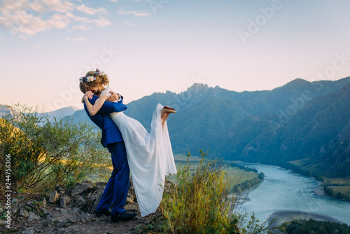 Young newly wed couple, bride and groom kissing, hugging on perfect view of mountains, river and blue sky