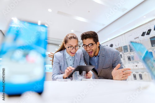 Cute multicultural couple in formal wear smiling and looking for new tablet to buy. Tech store interior.