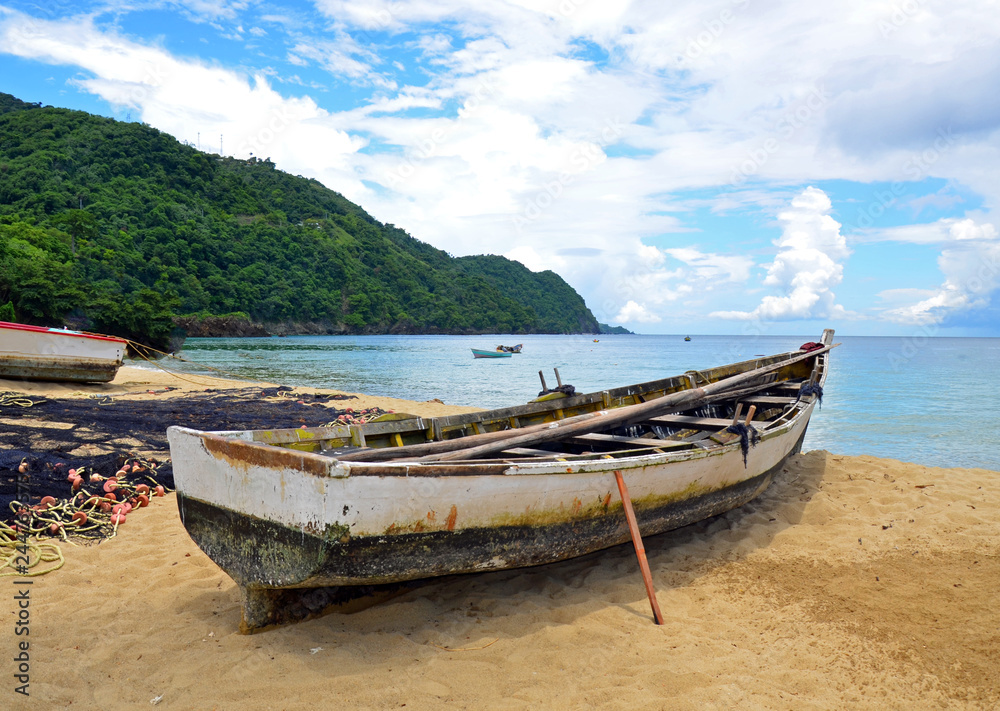 A rustic native Tobago wooden fishing boat propped up on a sandy beach by a drying net with orange floats and yellow rope, with others at anchor before lush rugged mountains under a cloudy blue sky. A rustic native Tobago wooden fishing boat propped up on a sandy beach by a drying net with orange floats and yellow rope, with others at anchor before lush rugged mountains under a cloudy blue sky.