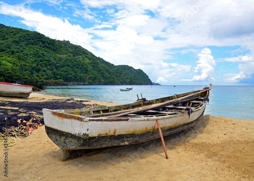 A rustic native Tobago wooden fishing boat propped up on a sandy beach by a drying net with orange floats and yellow rope  with others at anchor before lush rugged mountains under a cloudy blue sky.