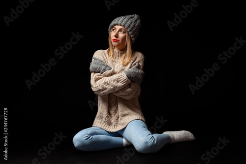 A blonde girl in gray knitted gloves, hat and sweater sitting on black floor with arms folded and hugging herself with her eyes looking up against a dark background alone, she is cold and lonely