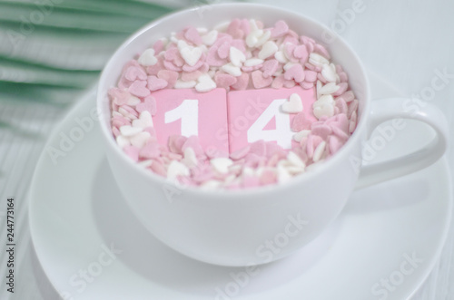 Pink square wood with white numbers 1 and 4.Gray square wood with white letters FEB.February 14 Valentine's Day.White coffee cup on wooden floor.Many small hearts, white and pink.