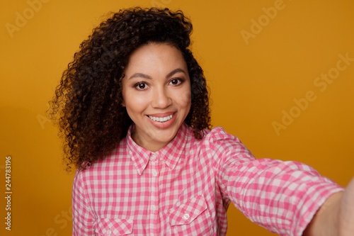 Excited black girl in cute yellow attire laughing while making selfie. Indoor portrait of gorgeous african female model taking picture of herself showing peace sign with fingers © burdun