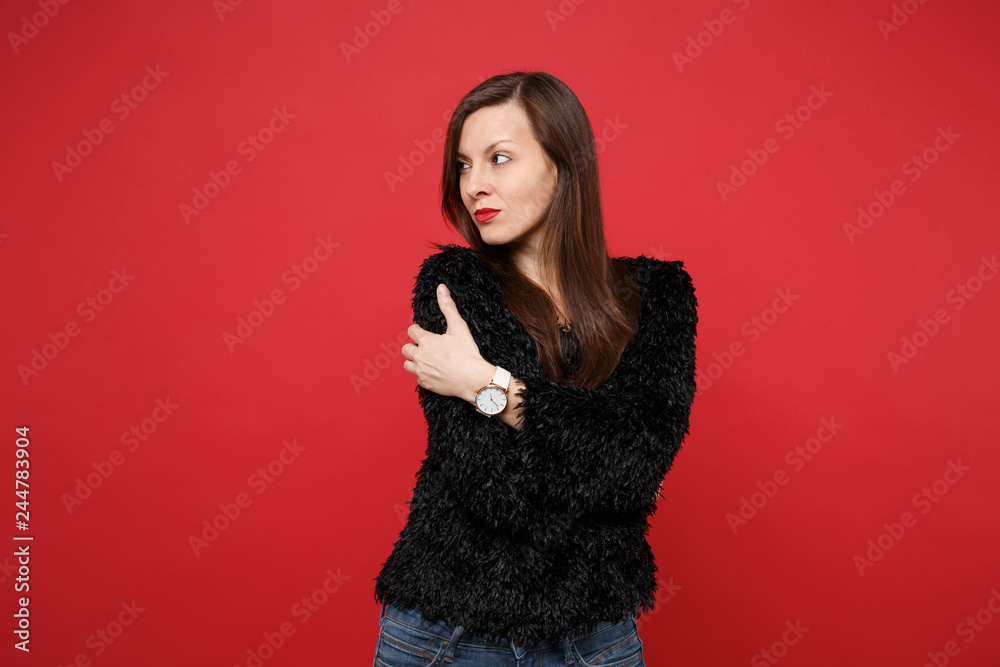 Fototapeta premium Portrait of pensive young woman in black fur sweater holding hands crossed, looking aside isolated on bright red background in studio. People sincere emotions, lifestyle concept. Mock up copy space.