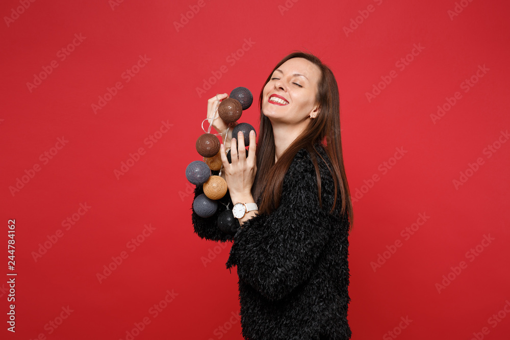Obraz premium Pretty young woman in black fur sweater keeping eyes closed holding round lights with garlands for decorating isolated on red background. People sincere emotions lifestyle concept. Mock up copy space.
