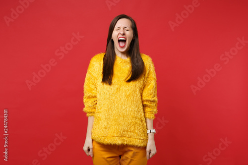 Portrait of weird crazy young woman in yellow fur sweater keeping eyes closed screaming isolated on bright red wall background in studio. People sincere emotions lifestyle concept. Mock up copy space.