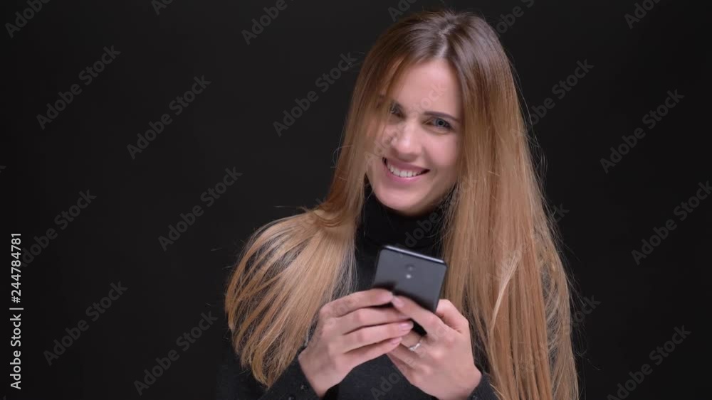 Portrait of young caucasian long-haired blonde girl smilingly watching into smartphone on black background. Portrait of young caucasian long-haired blonde girl smilingly watching into smartphone on black background.