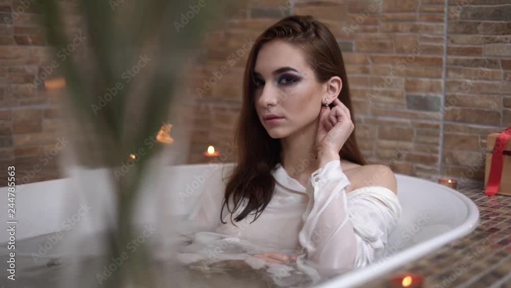 Beautiful sensual woman in white shirt taking a bath. Cute girl enjoying in the bathroom with burning candles.