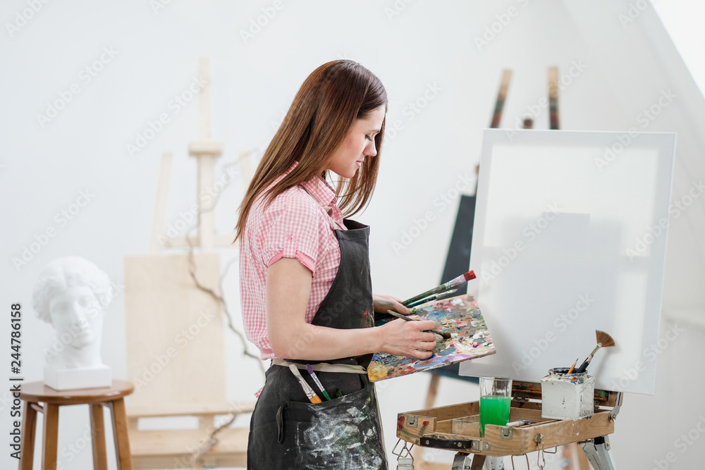 A young woman painter in a bright white studio draws a picture on canvas on an easel. A young woman painter in a bright white studio draws a picture on canvas on an easel.