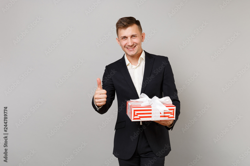 Joyful business man showing thumb up holding red striped present box with gift ribbon isolated on grey background. Achievement career wealth business. Valentine's Women's Day birthday holiday concept. Joyful business man showing thumb up holding red striped present box with gift ribbon isolated on grey background. Achievement career wealth business. Valentine's Women's Day birthday holiday concept.