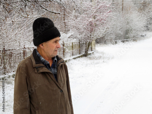 Elderly man standing on the rural street during a snowfall. Concept of cold weather, snow winter, homeless