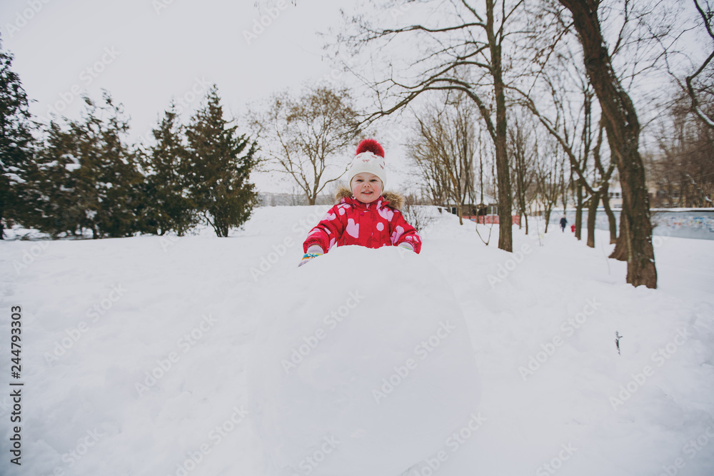 Cheerful little girl in winter warm clothes and hat playing with snowball, making snowman in snowy park or forest outdoors. Winter fun, leisure on holidays. Love relationship family lifestyle concept.