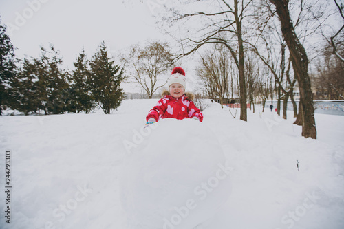 Cheerful little girl in winter warm clothes and hat playing with snowball, making snowman in snowy park or forest outdoors. Winter fun, leisure on holidays. Love relationship family lifestyle concept.
