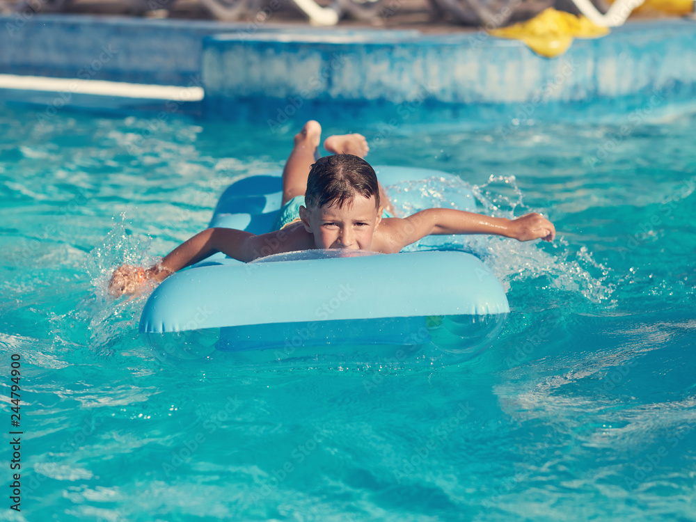 Smiling European boy is swimming on the inflatable blue floater in the hotel’s pool.