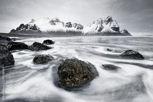 Snow-capped mountain formation reflected in the water of a black lava sand beach, in the foreground waves washed around by waves, great depth - Location: Iceland, south coast