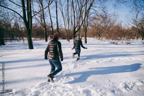 Back view of couple woman and man in warm clothes catching up with each other, walking in snowy park or forest outdoors. Winter fun, leisure on holidays. Love relationship people lifestyle concept.
