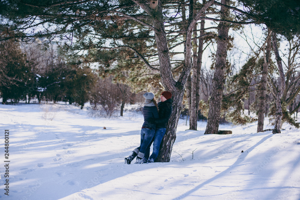Young couple woman, man in warm clothes hugging, looking at each other near tree, walking in snowy park or forest outdoors. Winter fun, leisure on holidays. Love relationship people lifestyle concept.