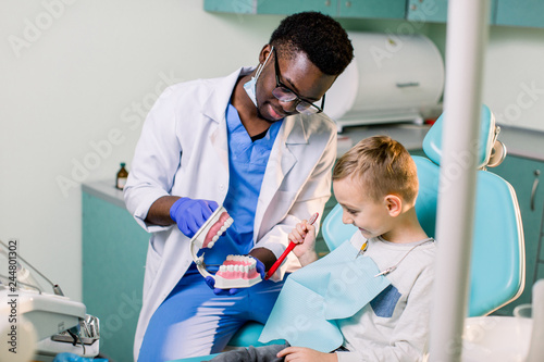 African dentist shows how to brush teeth to a small Caucasian boy a patient, using a artificial jaws. Dentistry, teeth hygiene concept