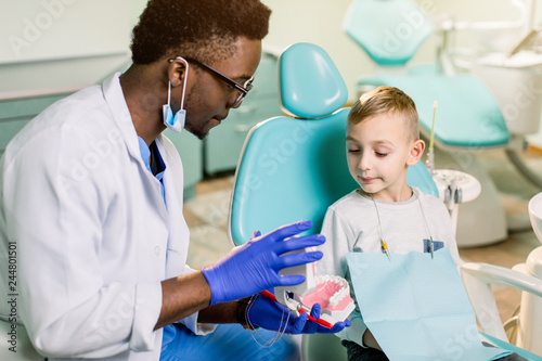 African American Man dentist in a uniform. Plastic jaw in the dentist s office. Dendist holds in his hands dental implants and talking to a little boy.