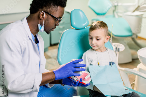 African American Man dentist in a uniform. Plastic jaw in the dentist s office. Dendist holds in his hands dental implants and talking to a little boy.