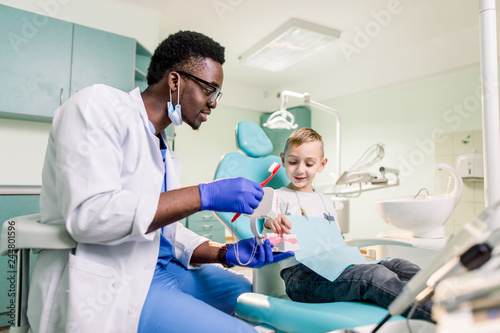Happy smiling black dentist tells Caucasian little boy how to brush his teeth. Caries prevention, Dentistry, teeth hygiene concept