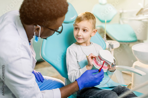 Little Caucasian boy is sitting in dental chair and smiling to African American dentist with artificial jaw in his hands