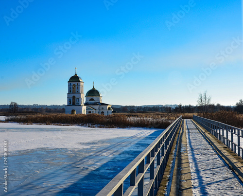 Snow-covered lake with a metal bridge towards church in the middle of a lake outdoor. Sunny day blue sky, clear weather