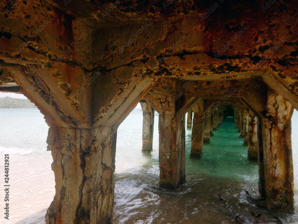 Looking through the shaded underside along the pilings of an old, rust covered, crumbling concrete pier that lies over sandy surf to aquamarine water that is washed out by the sunny day beyond.