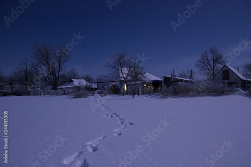 Night landscape of a snow-covered field. In the distance is seen the village. There are the footprints on the snow.