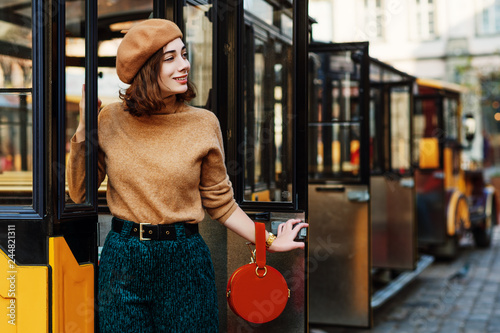 Outdoor portrait of young fashionable woman wearing beige beret, turtleneck, green corduroy trousers, belt, holding round orange suede handbag, posing in street of european city. Copy space for text