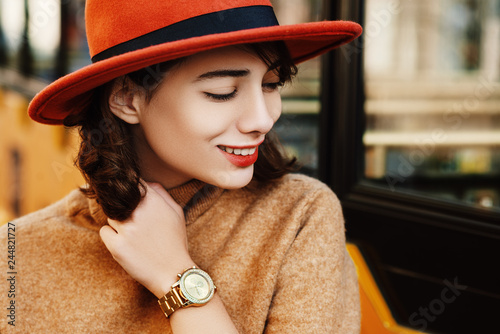 Close up outdoor portrait of young fashionable happy smiling woman wearing golden wrist watch, orange hat, beige turtleneck sweater. Copy, empty space for text