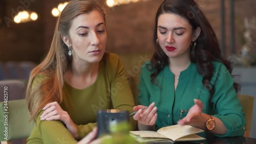 Two adorable young women are sitting in a cafe indoors and talking in a video chat with somebody by phone. One of them is asking something to clarify an answer and writing it down in her notebook. photo