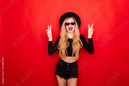 Young blonde woman in black hat and red sunglasses with peace gesture isolated on red background