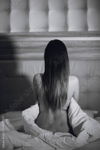 Back view of sexy young naked woman is sitting on white bed. Black and white photo.