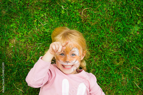 beautiful little girl at the children's party with a butterfly pattern on her face, posing emotionally while lying on the bright green grass