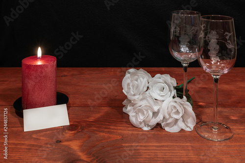 Beautiful etched wine glasses with awhite roses and red candle and name tag on wooden table and dark background.