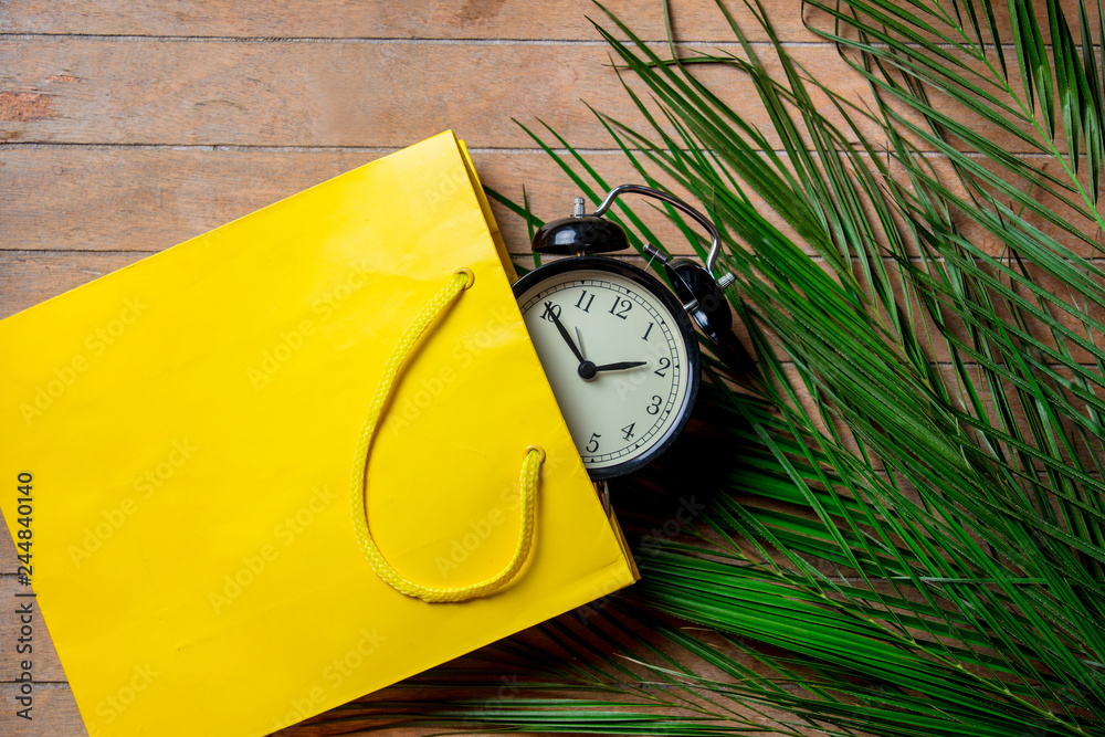 Fototapeta premium Vintage alarm clock in yellow bag and palm leaves on wooden table. Above view