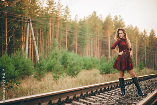 Beautiful dreamy girl with curly natural hair enjoy nature in forest on railway. Dreamer lady in burgundy dress walk on railroad. Inspired girl on rails at dawn. Sun in hair in autumn. Good mood.