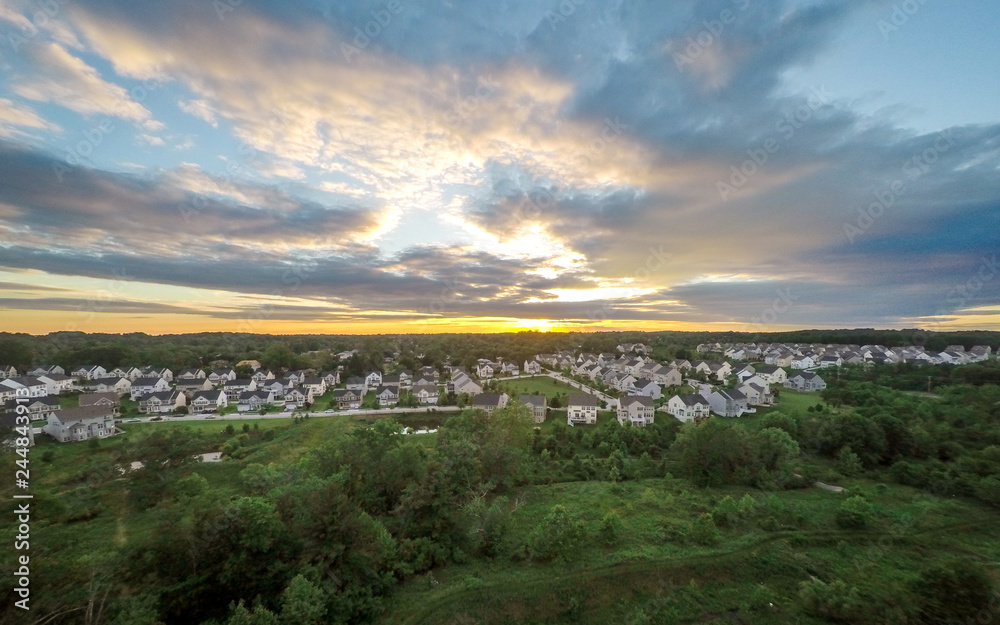 Vibrant, Aerial Drone Photo of a Suburban Neighborhood at Sunset - with a Yellow Horizon, Green Trees, Thick Clouds and a Blue Sky with a Reservoir in the Foreground