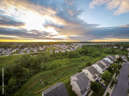 Vibrant, Aerial Drone Photo of Rows of Houses in the Suburbs at Sunset - with a Yellow Horizon, Green Trees, Thick Clouds, a Blue Sky, a Reservoir and More Homes in the Background © Jon