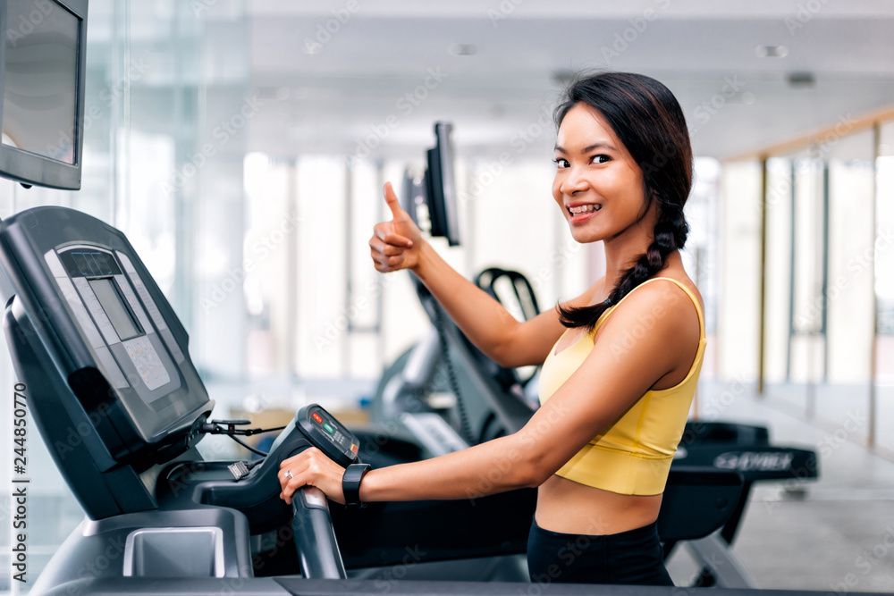 Attractive Asian woman smiling and running on a running treadmill at gym. Fitness model giving thumbs up Attractive Asian woman smiling and running on a running treadmill at gym. Fitness model giving thumbs up