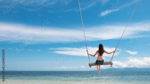 Young beautiful happy woman in top and shorts swinging on a swing on the shore of a tropical sea during vacation. Travel and vacation concept