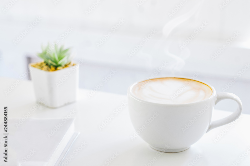 White cup of Latte art coffee as smoke with Flower Pot on wood table in the morning at home office,Clean white interior White cup of Latte art coffee as smoke with Flower Pot on wood table in the morning at home office,Clean white interior