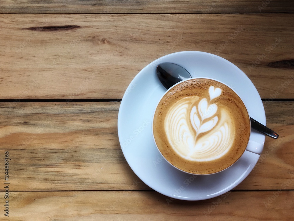 Hot cappuccino coffee in white cup and saucer with spoon on wooden table background. Art of milk foam drawing like white heart on brown melt coffee top view Hot cappuccino coffee in white cup and saucer with spoon on wooden table background. Art of milk foam drawing like white heart on brown melt coffee top view